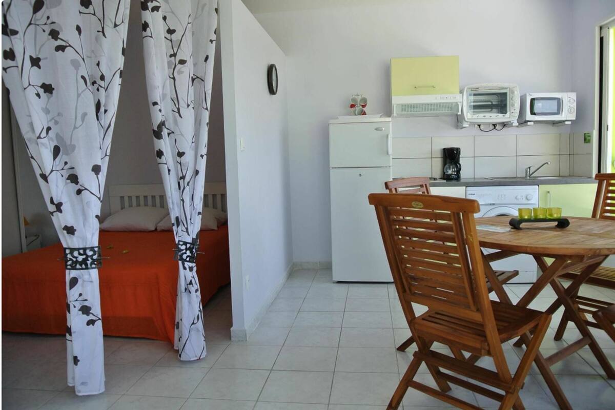 A bedroom with a bed, a window with curtains, a kitchenette with a fridge, microwave, and toaster, and a dining area with a table and chairs.