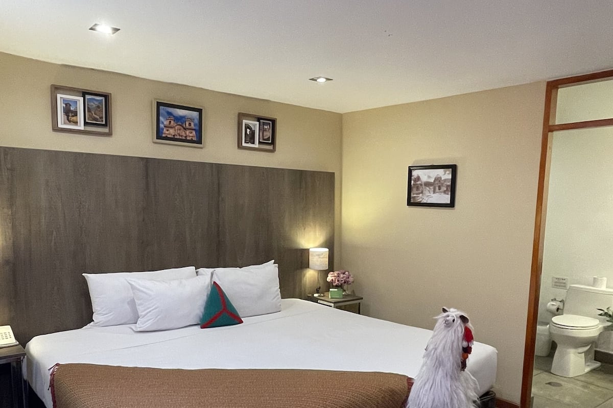 A hotel room with a large bed, a bedside table with a lamp, a small bench, and a wall-mounted picture.