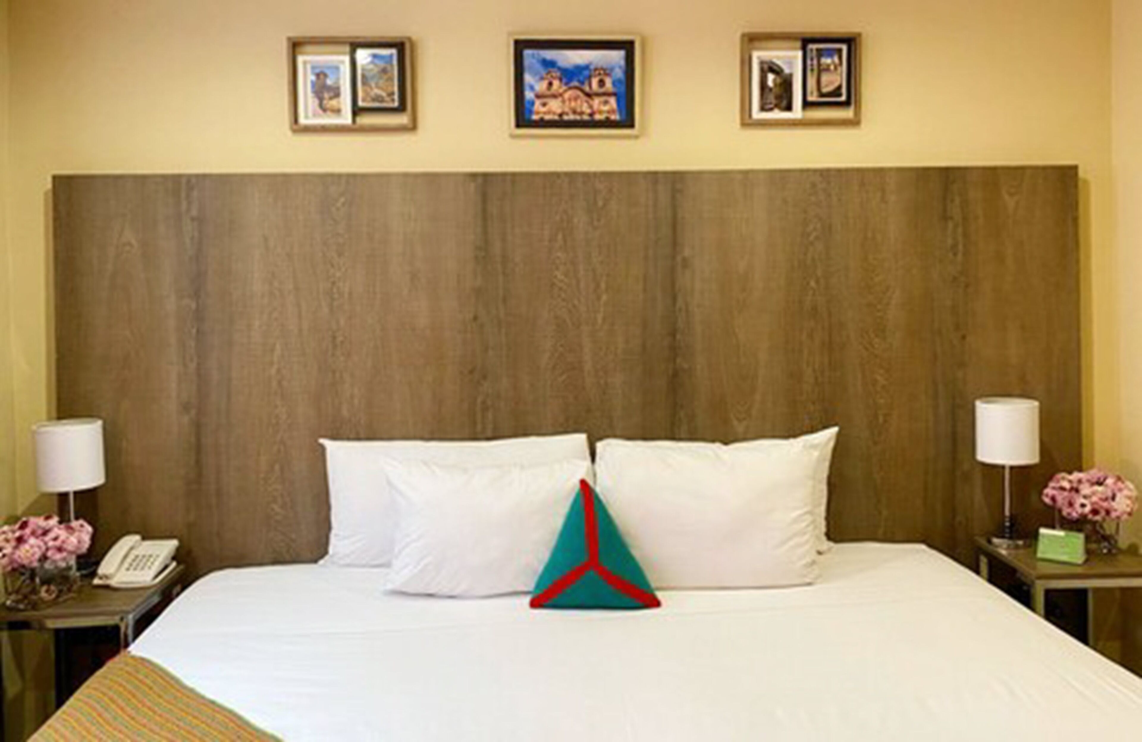 Ein Hotelzimmer mit einem hölzernen Kopfteil, einem Bett mit weißer Bettwäsche, zwei Nachttischen mit Lampen und einem Telefon sowie drei gerahmten Bildern an der Wand.
