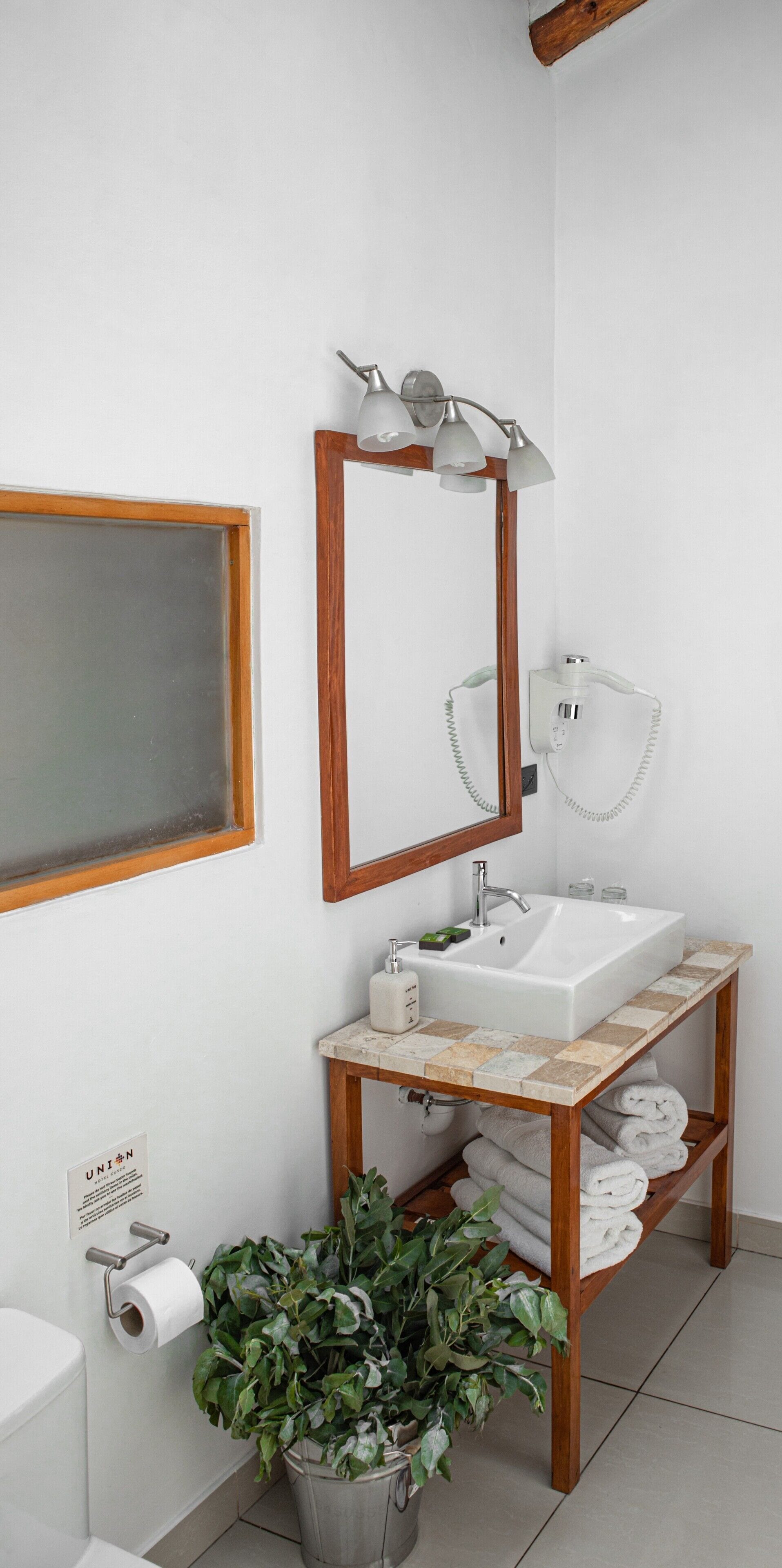 Een badkamer met een wit wastafel, houten wastafelmeubel, een spiegel met verlichting, een toilet en een potplant.