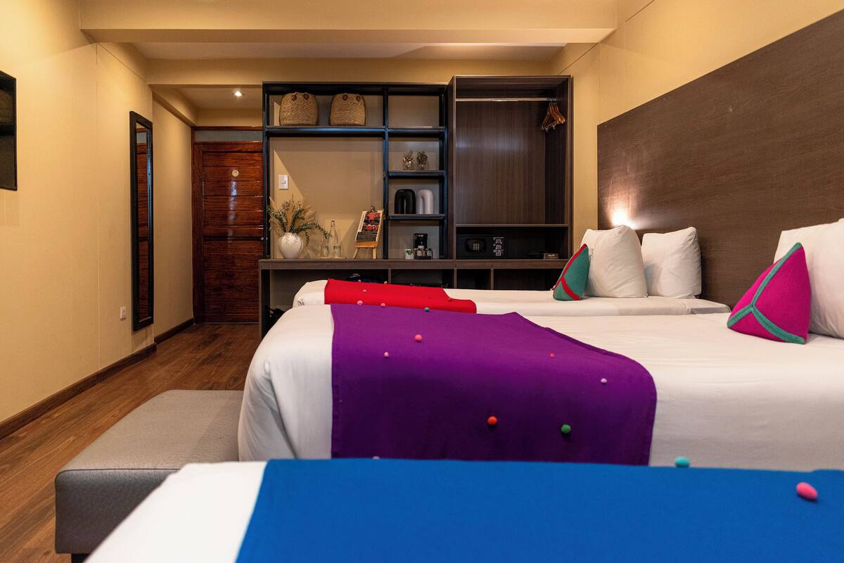 A hotel room with a bed, a seating area, a wooden wardrobe, and a shelf with various items.