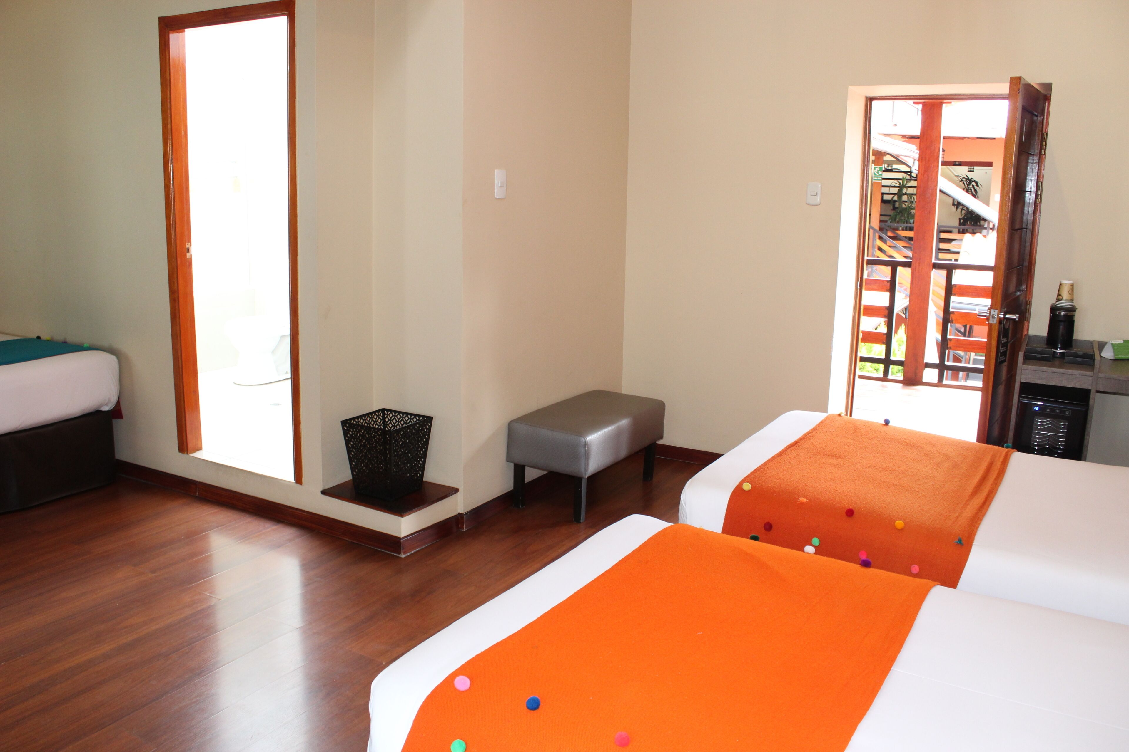 A hotel room with two beds, a small table, a bench, and a door leading to a balcony.