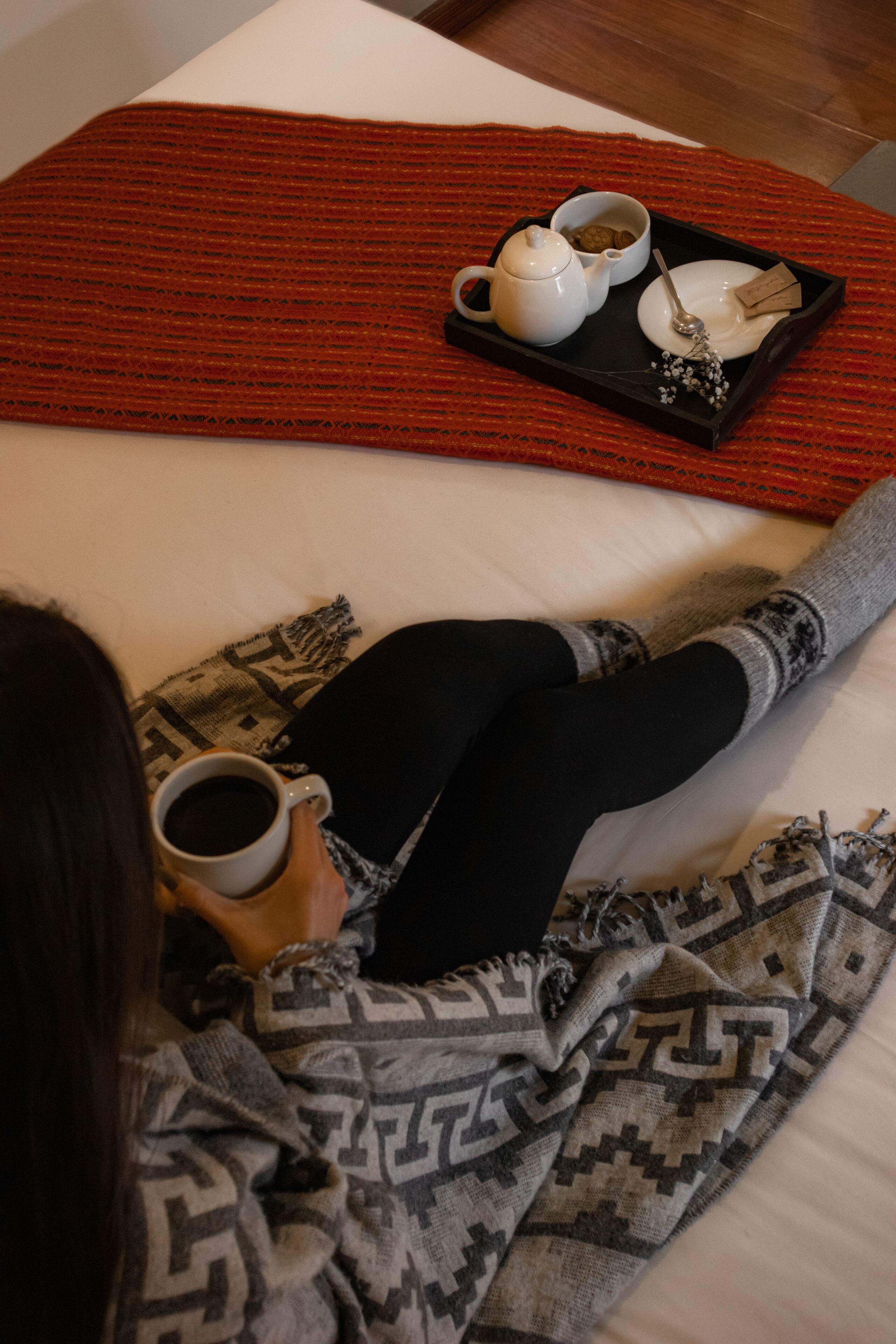 Una persona estesa en un llit amb una safata que conté una teiera, una tassa, una cullera i una xícara de cafè.