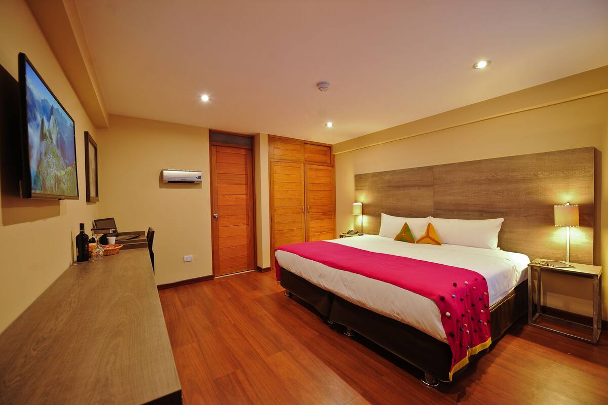 A hotel room with a large bed, wooden wardrobe, a desk, and a television on the wall.