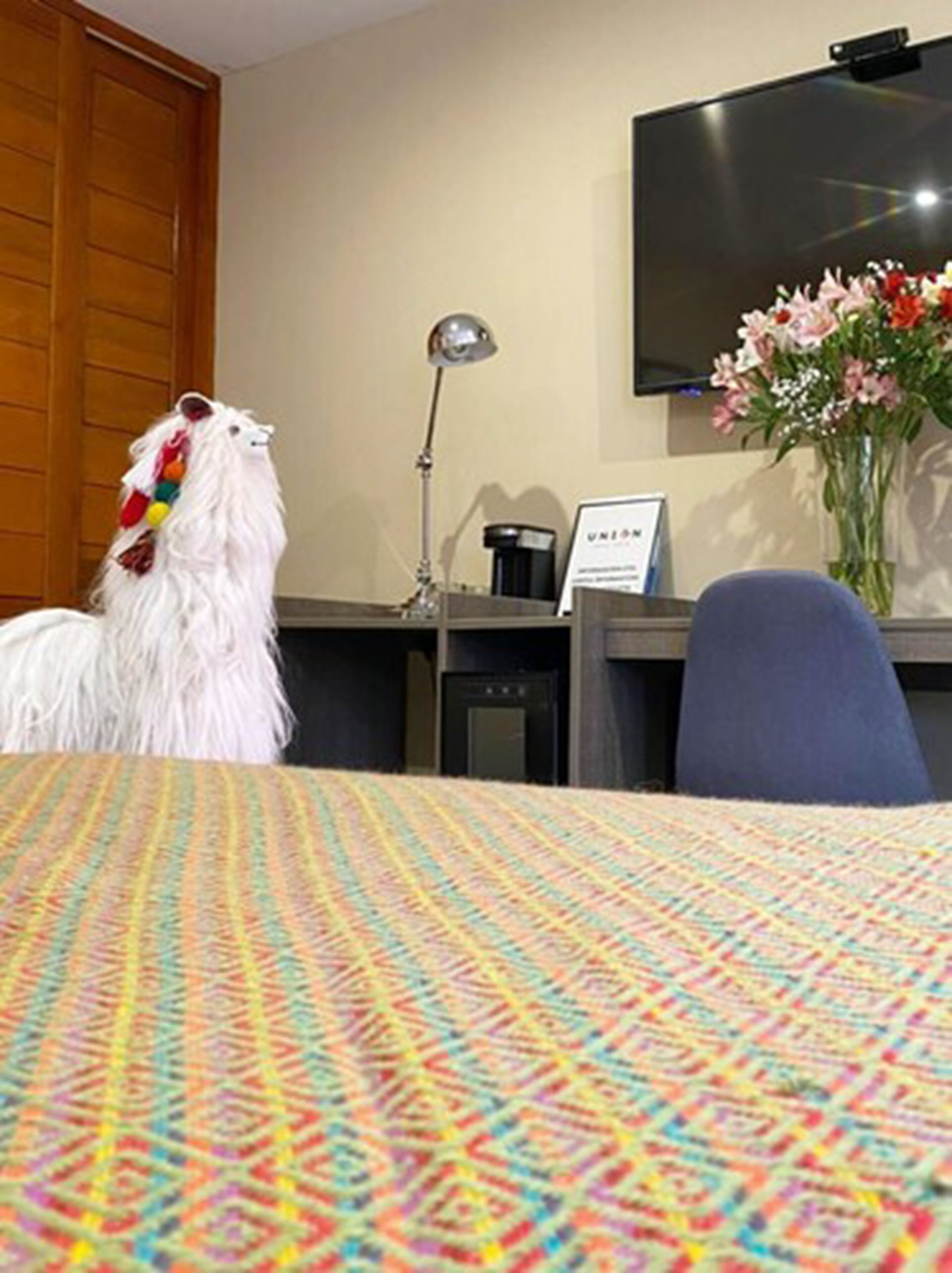 A hotel room with a bed, a desk, a chair, a lamp, a TV, and a vase with flowers.