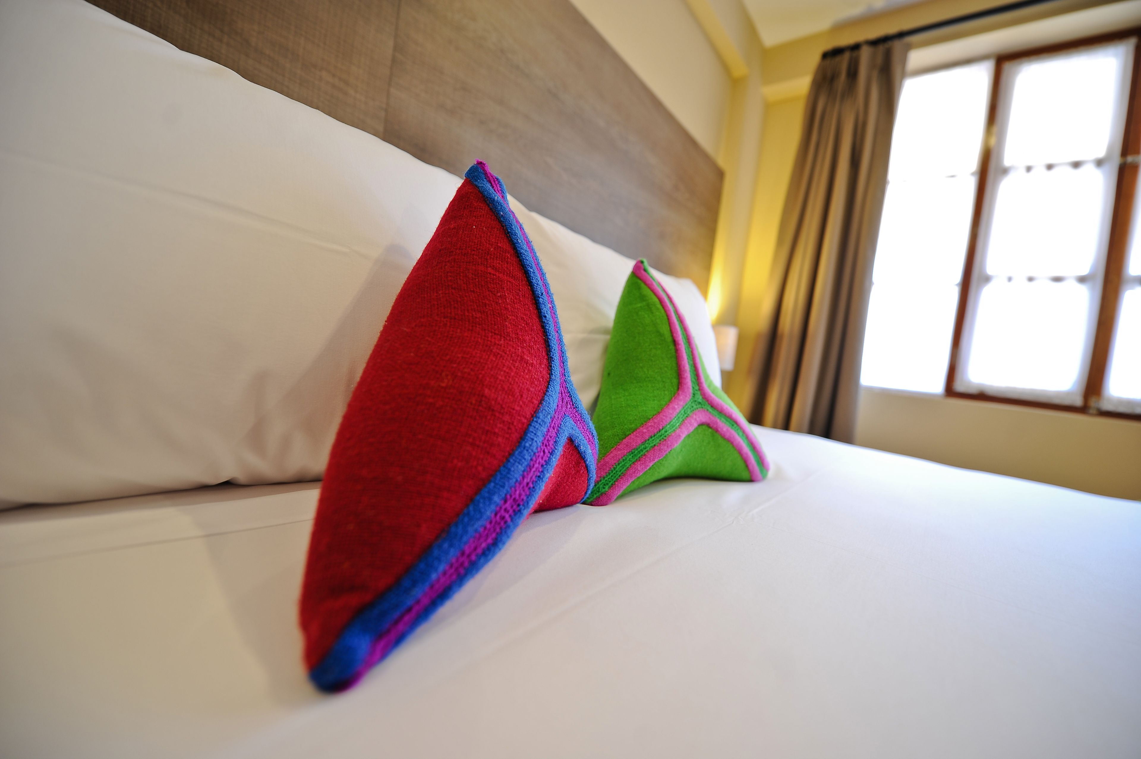 Uma cama com lençóis brancos e almofadas coloridas em quarto com cabeceira de madeira e janela com cortinas.