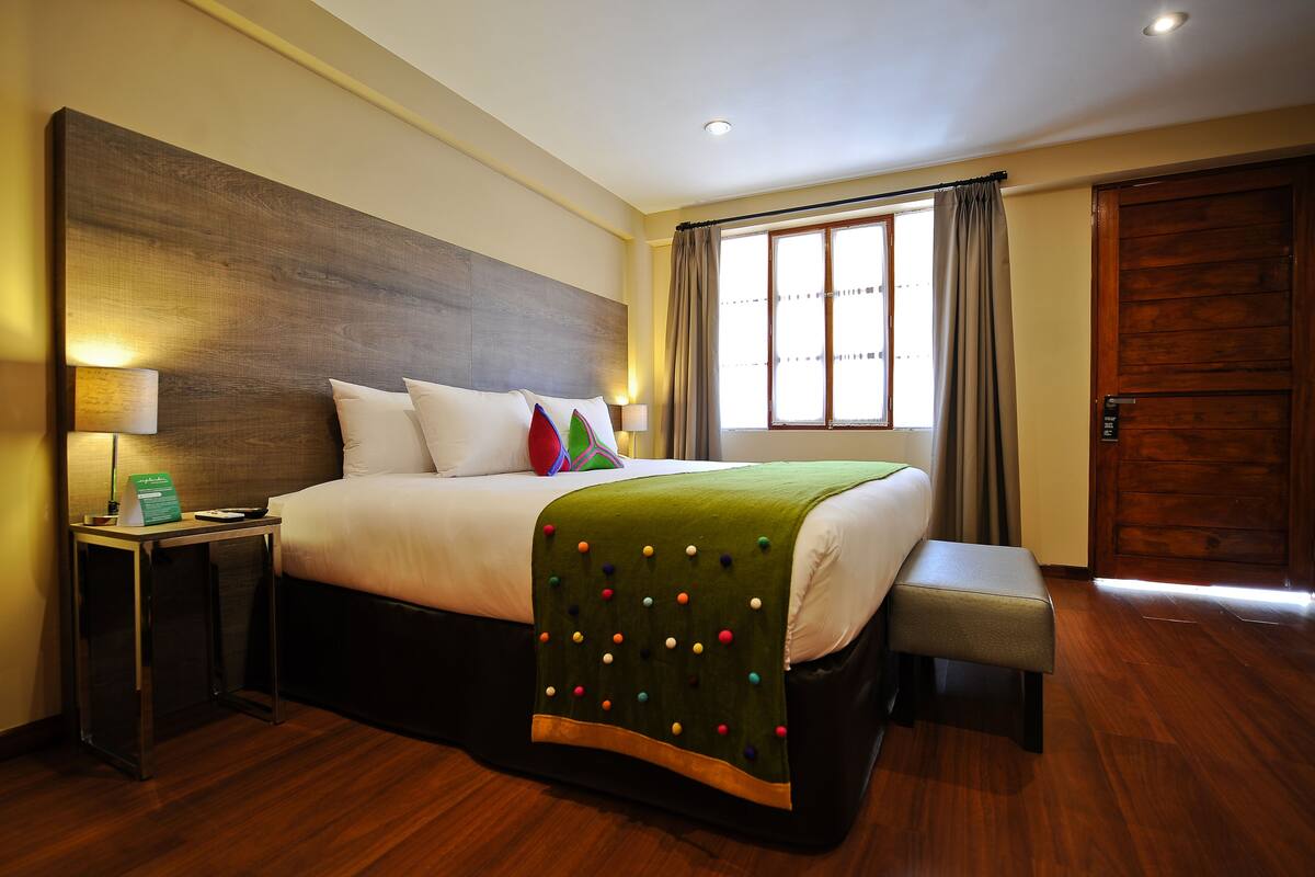 A hotel room with a large bed, a bedside table with a lamp, a bench, and a window with curtains.