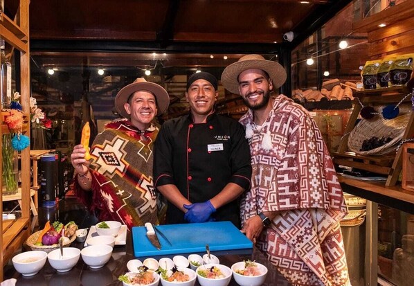 Three people standing in a restaurant with a variety of dishes and a blue cutting board on the counter.