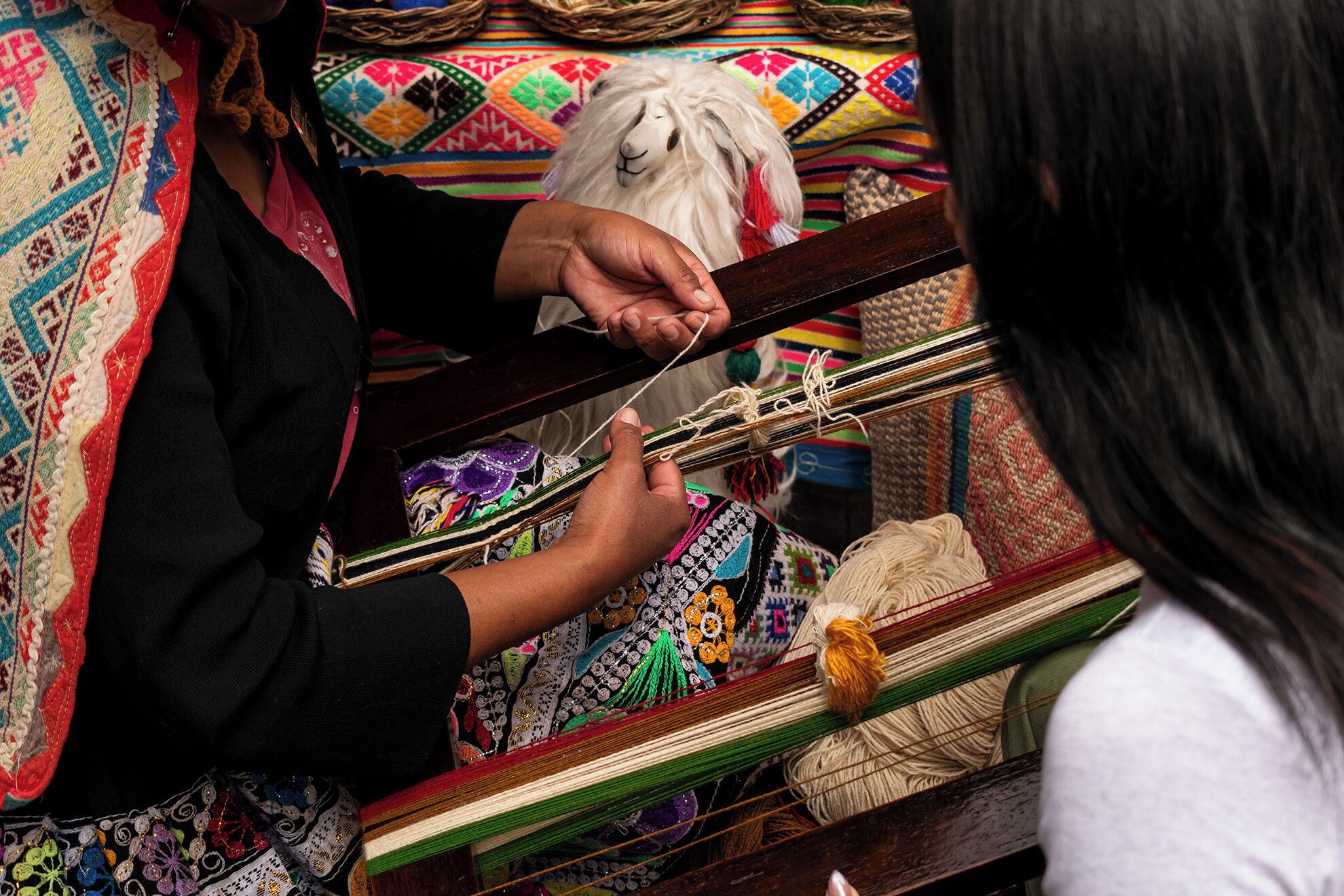 A person weaving on a loom with colorful textiles and a white sheep in the background.