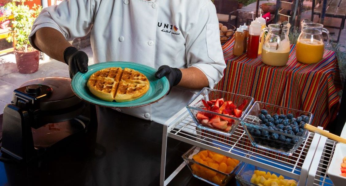 A chef holding a plate of waffles, with a breakfast buffet table in the background.