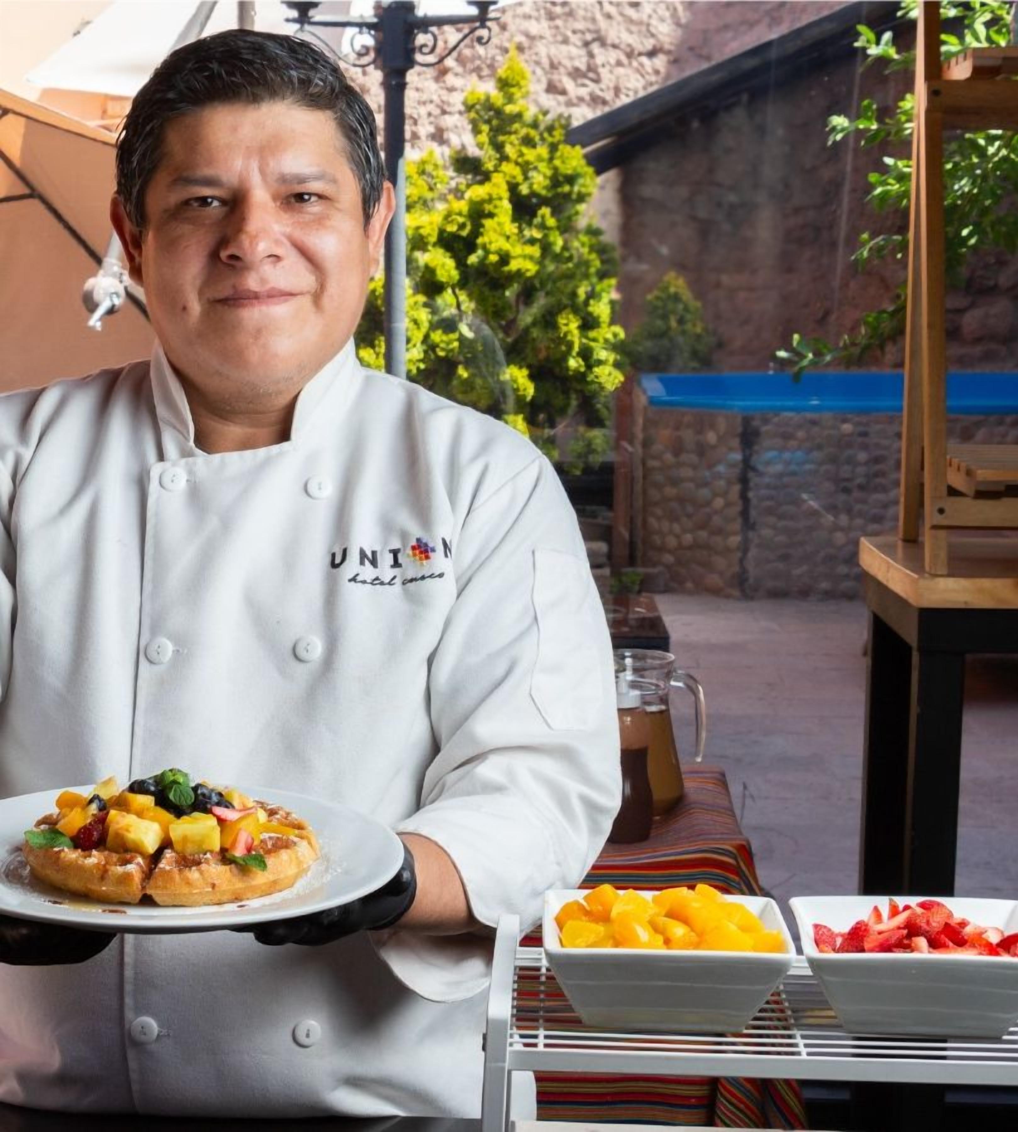 A chef in a white uniform holding a plate of food, with a sign reading "Breakfast Cooking Show All Inclusive" in the background.