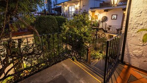 Edificio residencial con una escalera que lleva a un balcón, rodeado de follaje y un muro de piedra.