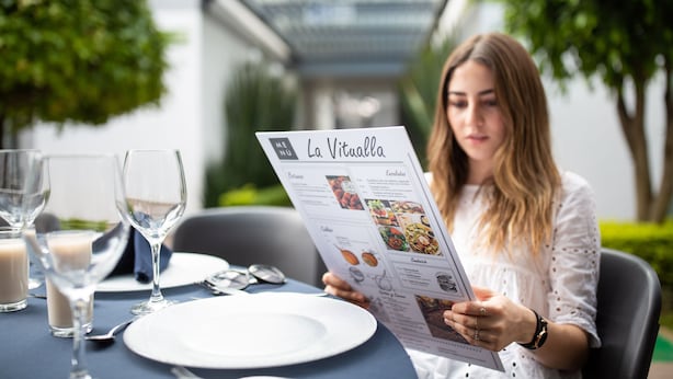 A person reading a menu at an outdoor dining table with a glass of white wine and a glass of milk. - Hotel Horizon and Convention Center (Morelia)