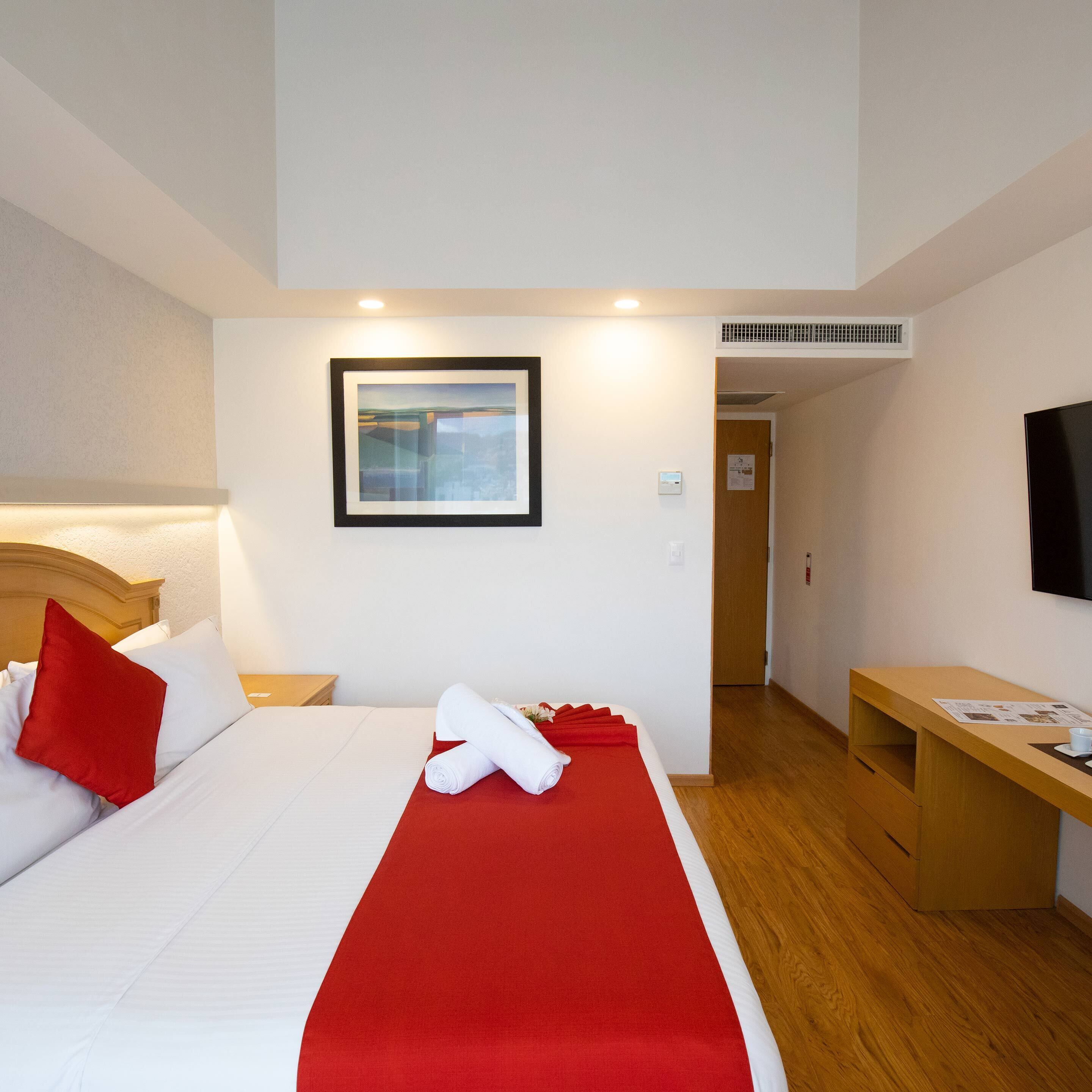 Een hotelkamer met een bed, een bureau, een televisie en een ingelijst kunstwerk aan de muur.