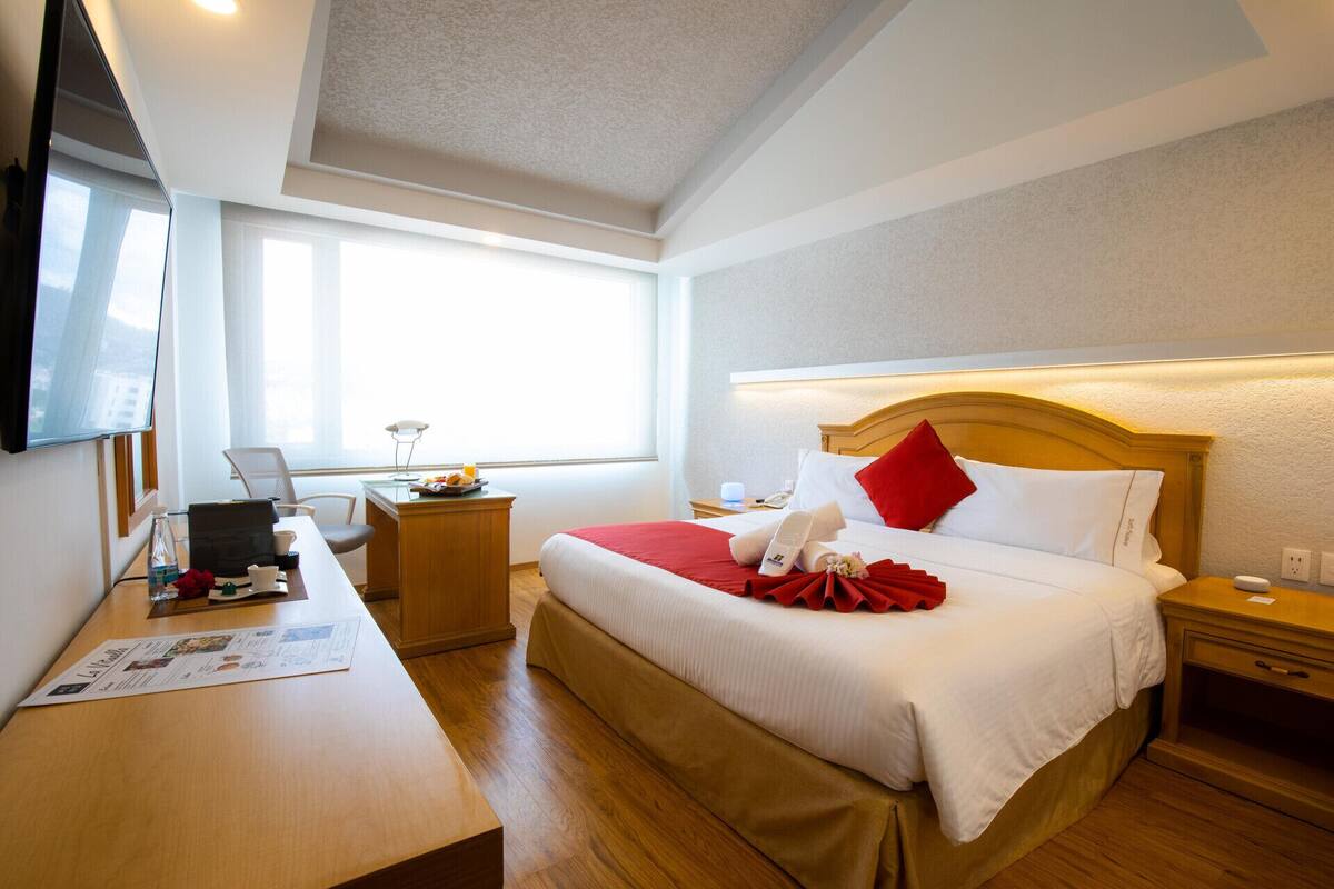 A hotel room with a bed, bedside table, lamp, and a desk with a laptop and papers.
