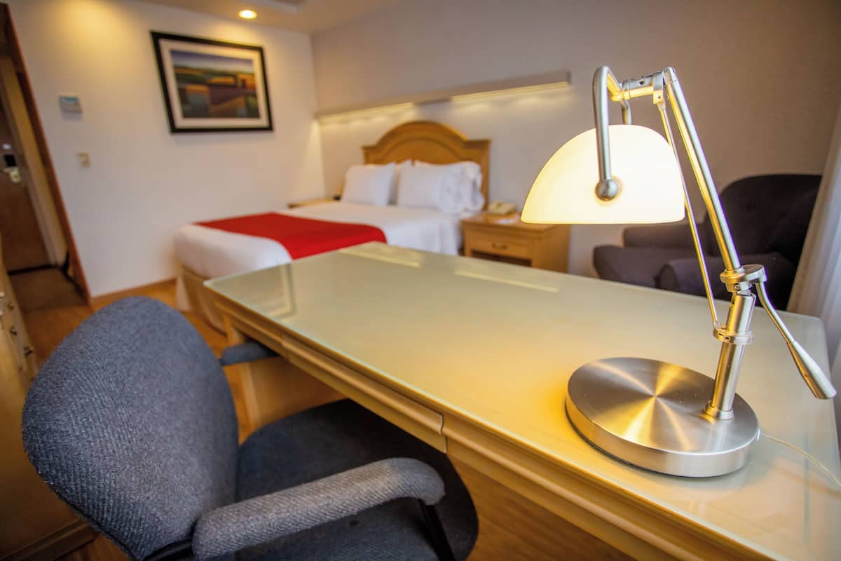 A hotel room with a bed, bedside table, lamp, chair, and a painting on the wall.