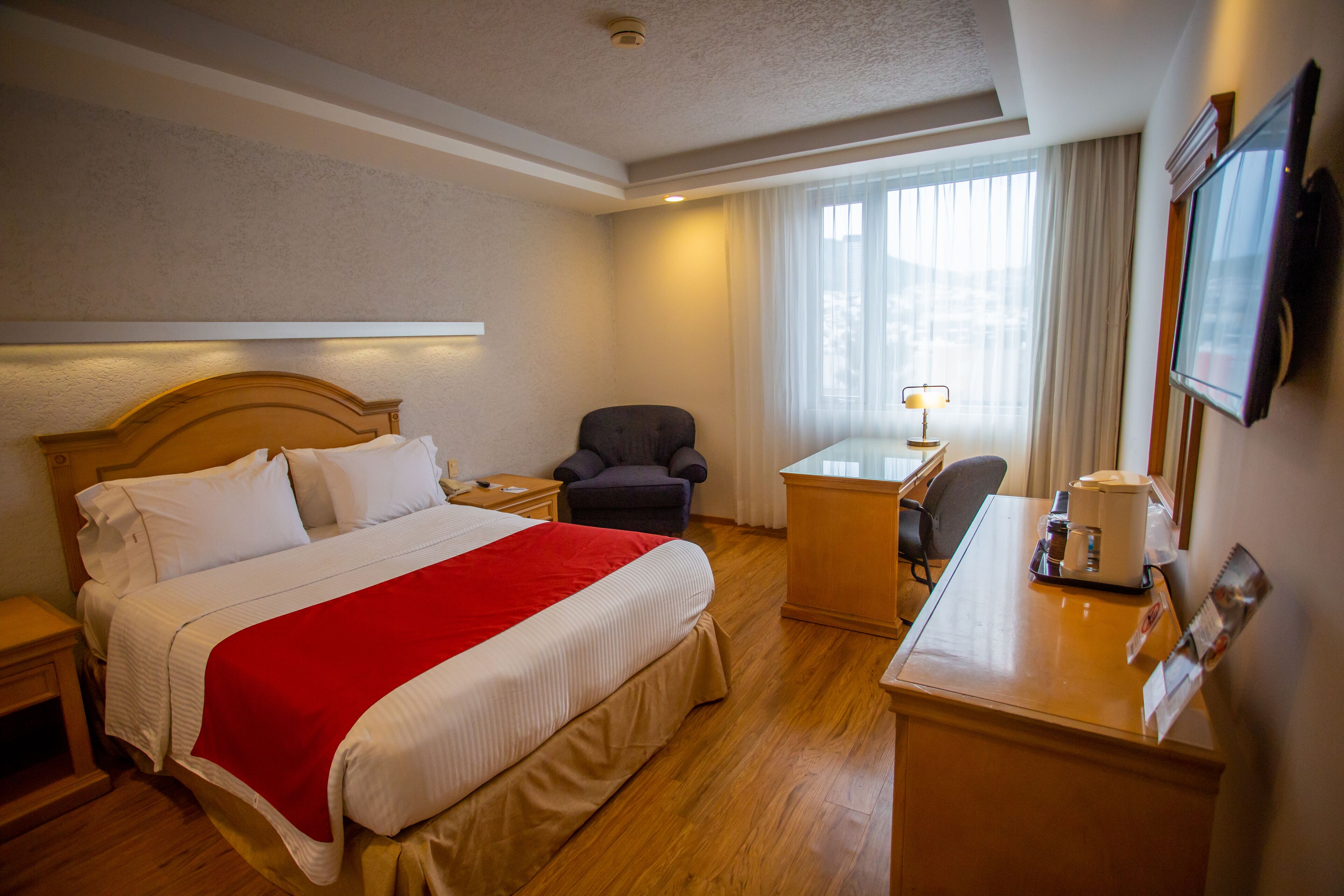Una habitación de hotel con una cama grande, un escritorio, una silla, un televisor y una ventana con cortinas.