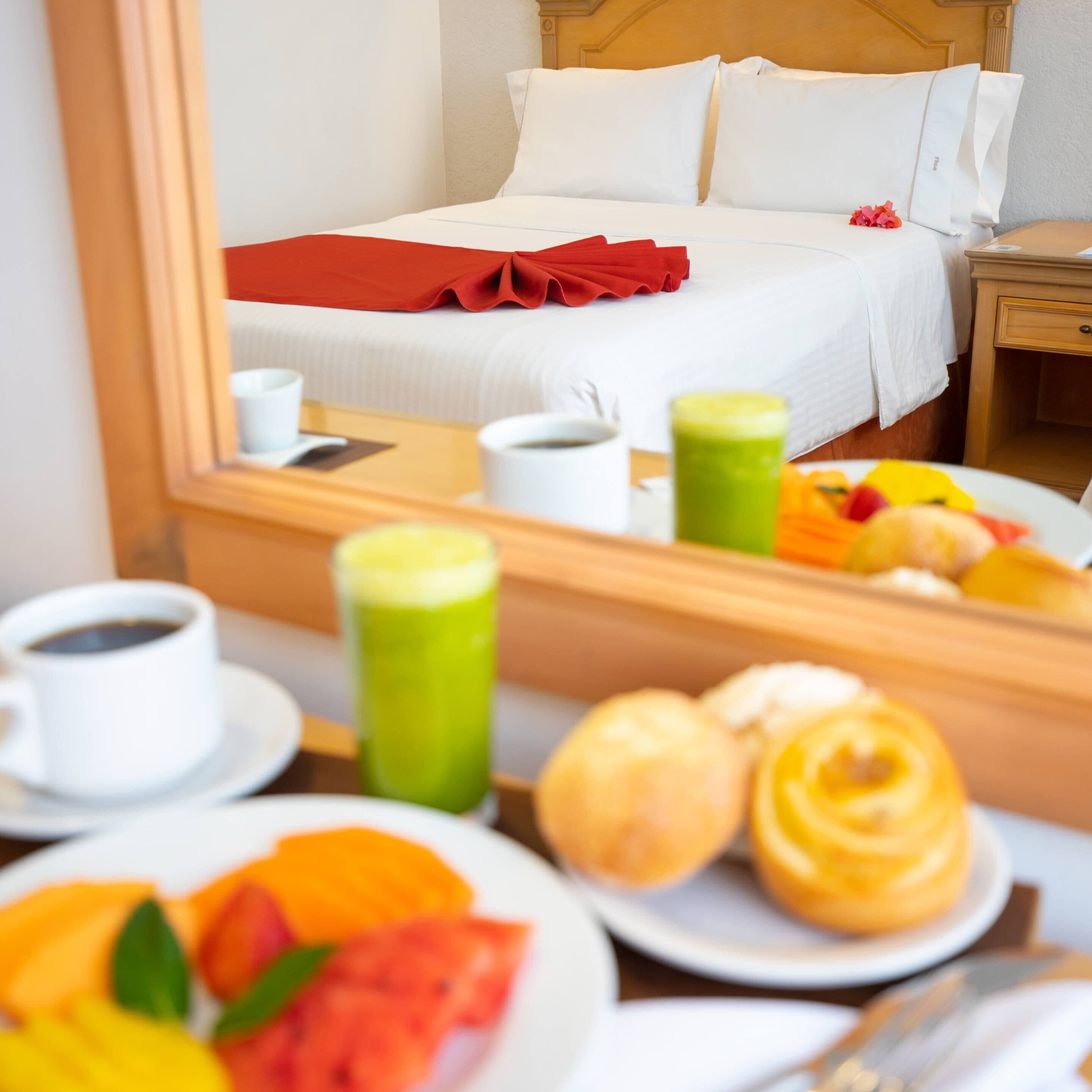 A hotel room with a bed, a bedside table with a lamp, and a breakfast tray with fruit and pastries.