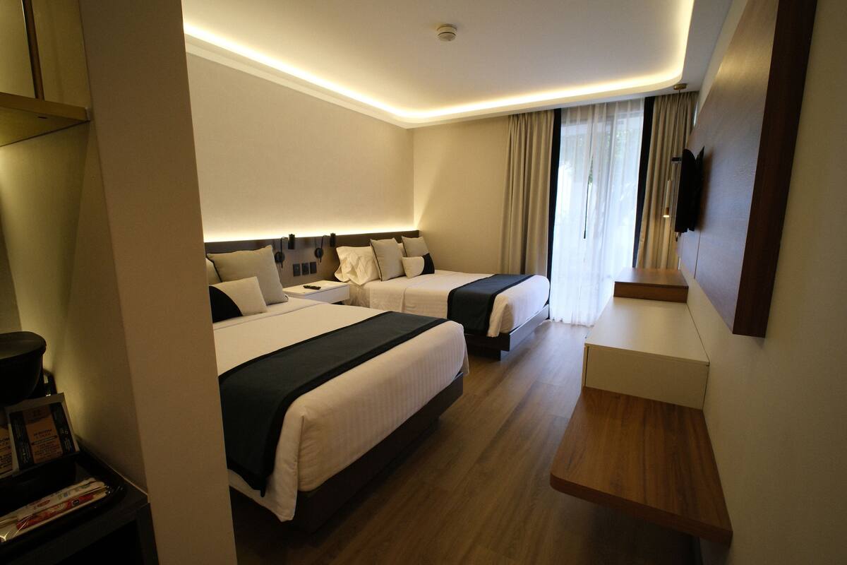 A hotel room with two beds, a bedside table, a TV, and a wooden bench.