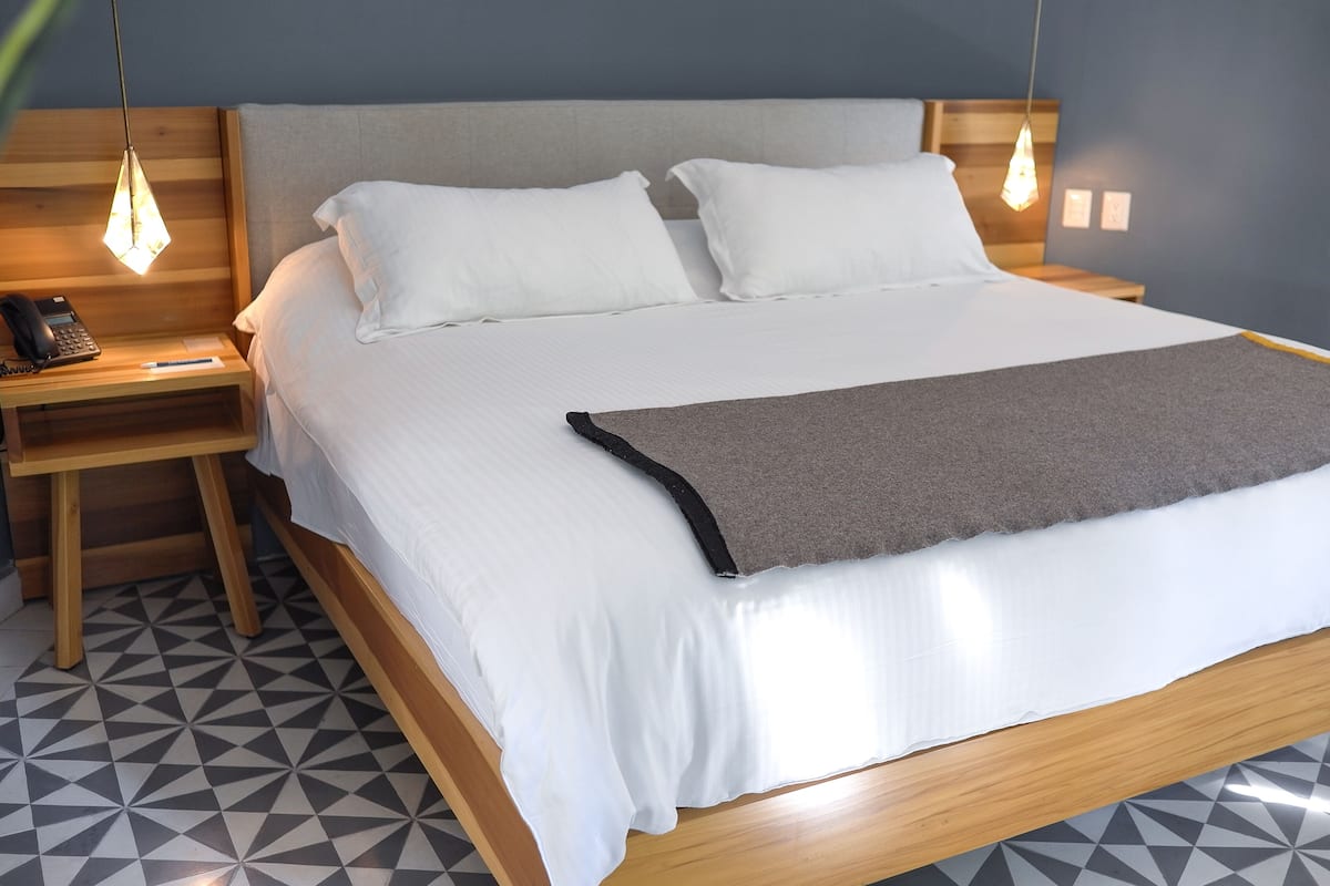 Una cama bien tendida con sábanas blancas y una manta gris, cabecero de madera, mesitas de noche con lámparas y un suelo con estampado geométrico.