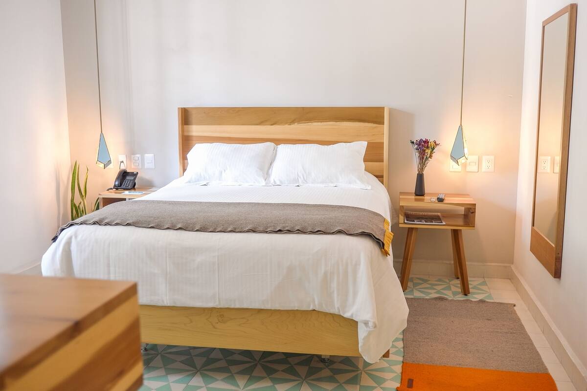 Un dormitorio ordenado con cama de madera, ropa de cama blanca, una mesita de noche con lámpara y un pequeño escritorio con un jarrón de flores.