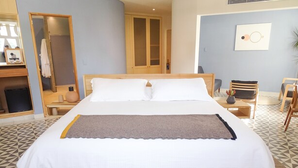 Una cama bien hecha con cabecera de madera, una mesita de noche con lámpara y una silla.