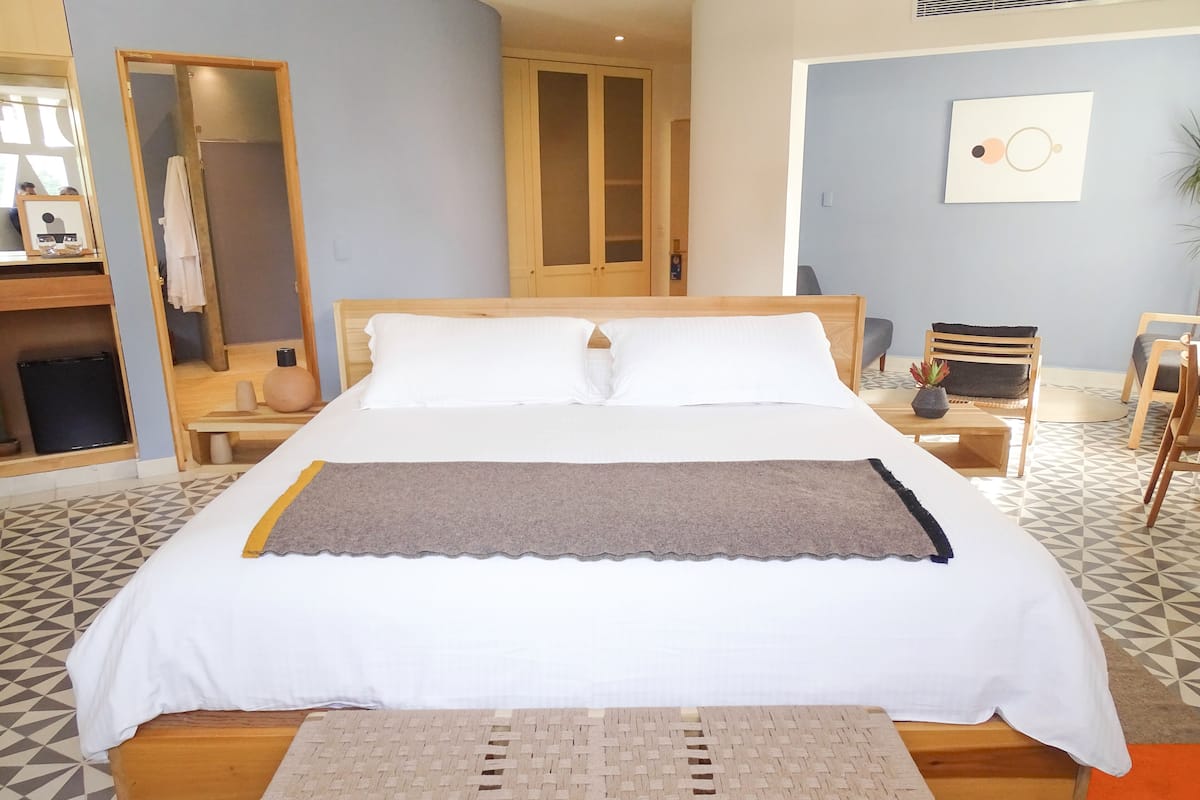 Una cama bien hecha con cabecera de madera, una mesita de noche con lámpara y una silla.