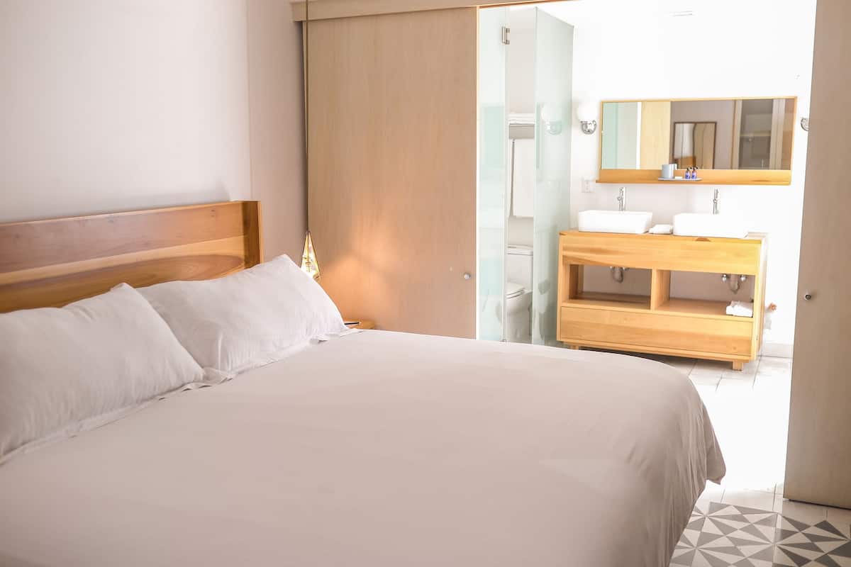 Habitación de hotel con cama de madera, ropa de cama blanca, tocador de madera con espejo y piso de baldosas.