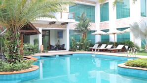 A swimming pool with lounge chairs and umbrellas, surrounded by tropical plants and a modern building with large windows. - Palm Spring Resort & Spa (Yangon)