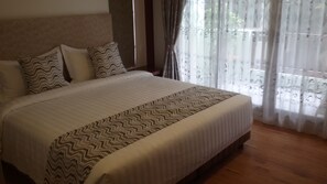 A neatly made bed with a patterned bedspread and pillows, a wooden headboard, and sheer curtains with a floral pattern.