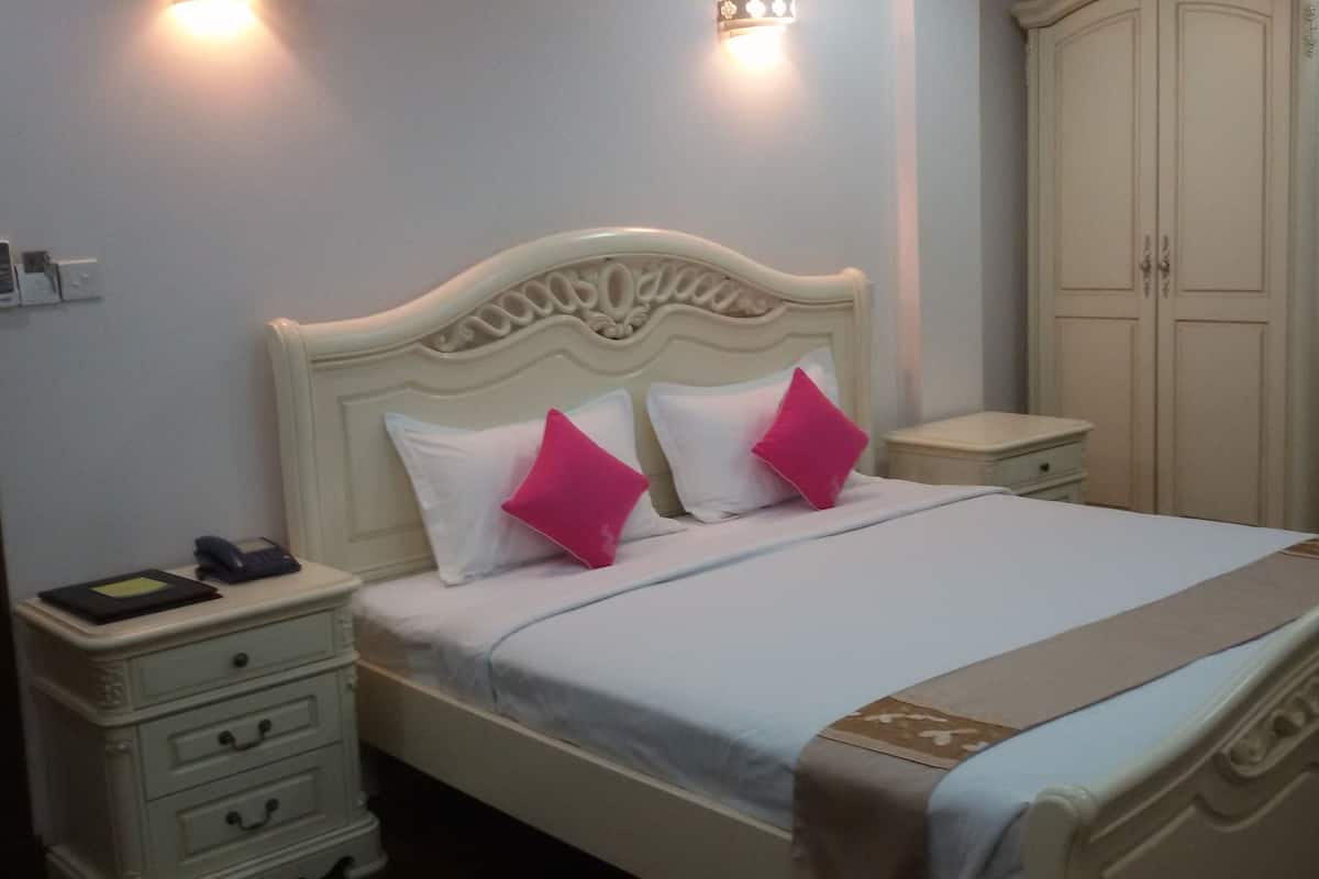 A neatly made bed with a white headboard, two pink pillows, and a light blue bedspread. There are two bedside tables with drawers, a nightstand, and a wardrobe in the background.