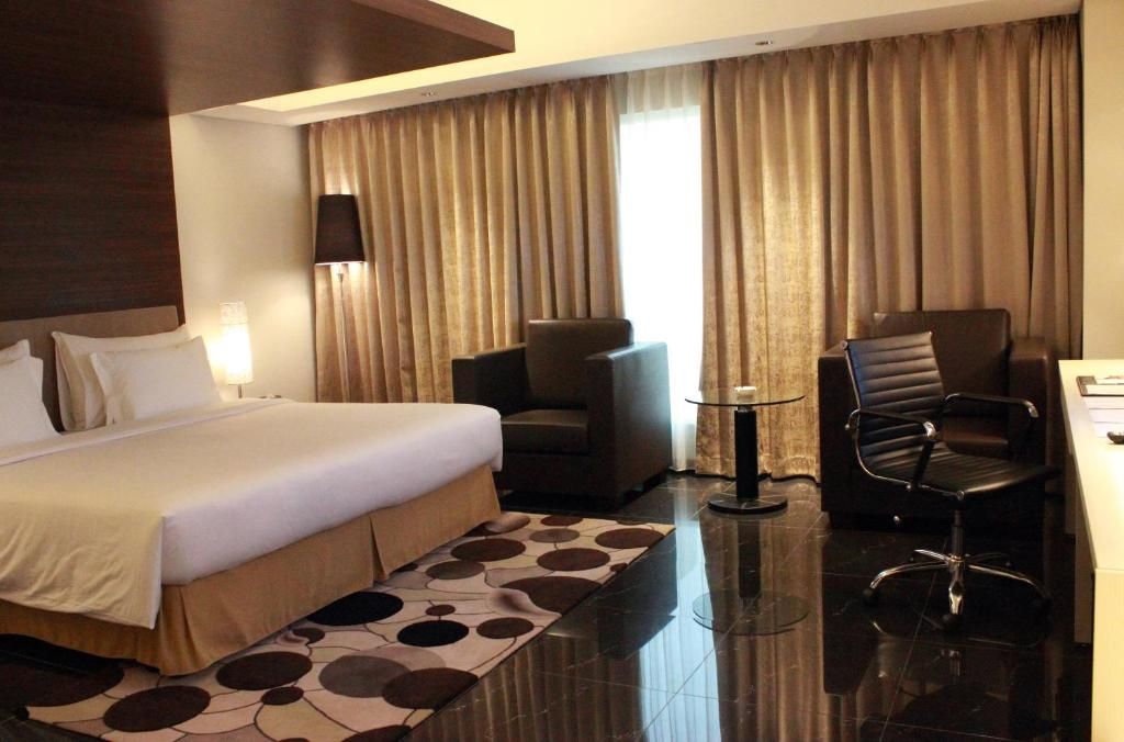 A hotel room with a bed, two chairs, a desk, and a large window with curtains.