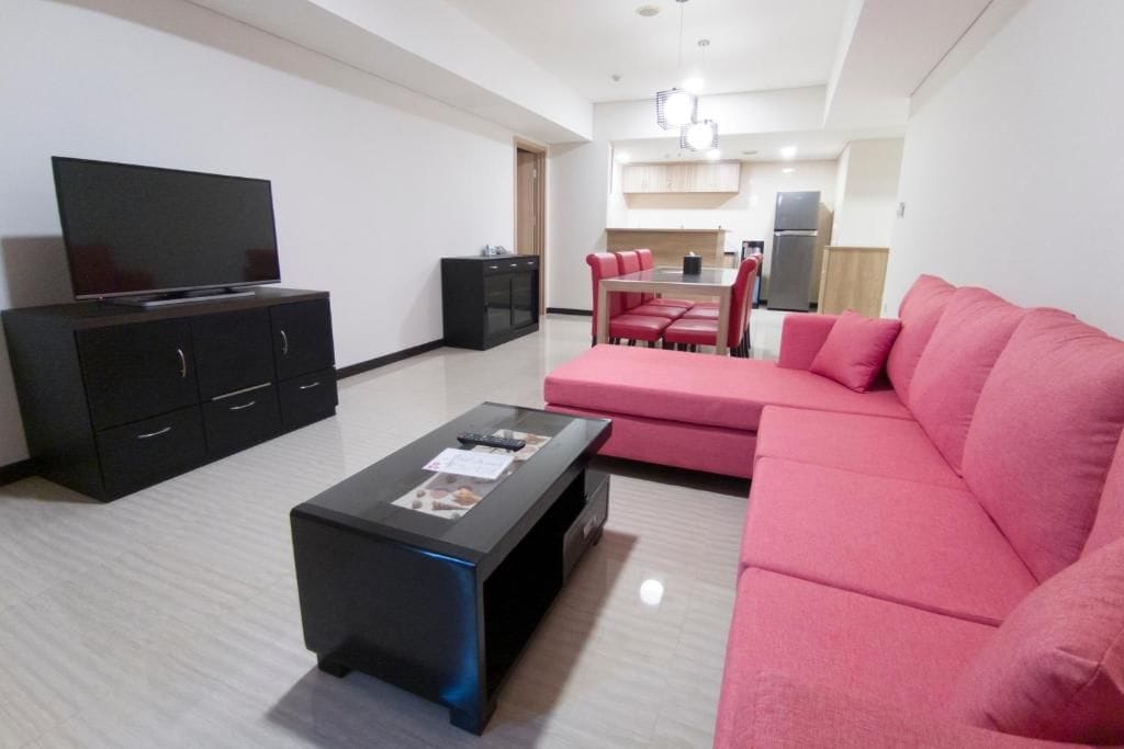 A living room with a flat-screen TV, a black coffee table, a pink sofa, and a dining area with a table and chairs.