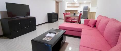 A living room with a flat-screen TV, a black coffee table, a pink sofa, and a dining area with a table and chairs.