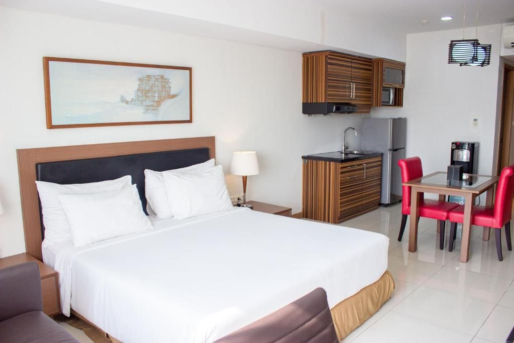 A hotel room with a bed, a desk, a chair, a kitchenette, and a painting on the wall.