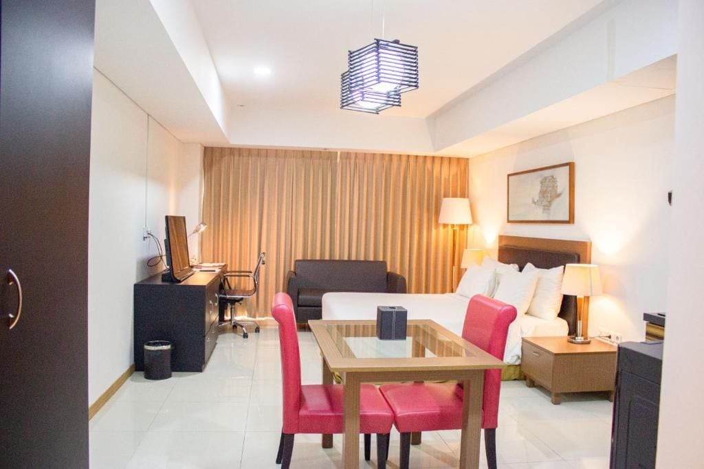 A hotel room with a bed, bedside table, lamp, desk with computer, chair, and a hanging light fixture.
