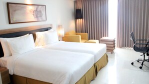 Une chambre d’hôtel avec deux lits, un bureau, une chaise, une lampe et une grande fenêtre avec des rideaux.