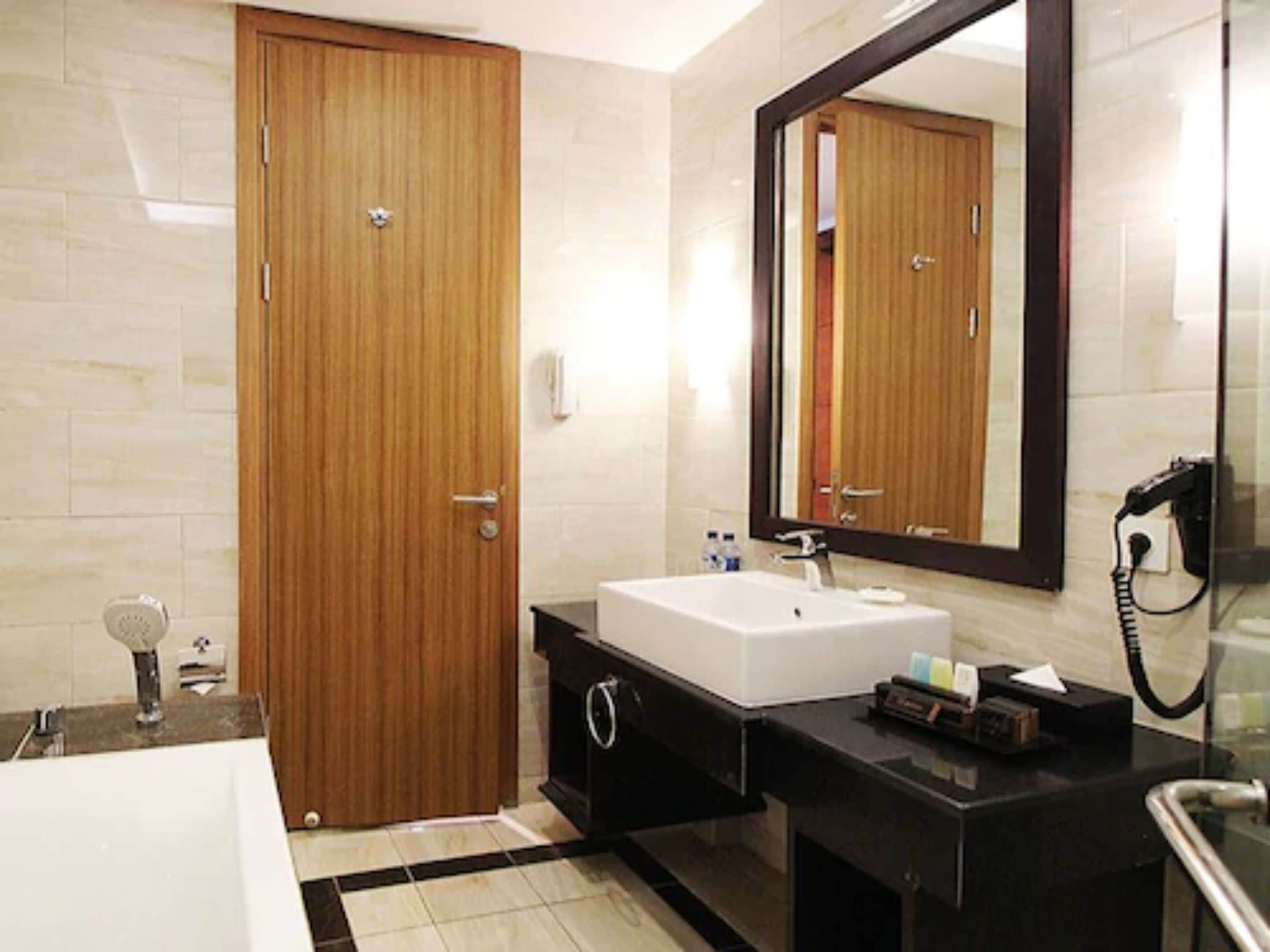 Un baño moderno con puerta de madera, lavamanos blanco, espejo grande y cabina de ducha de vidrio.