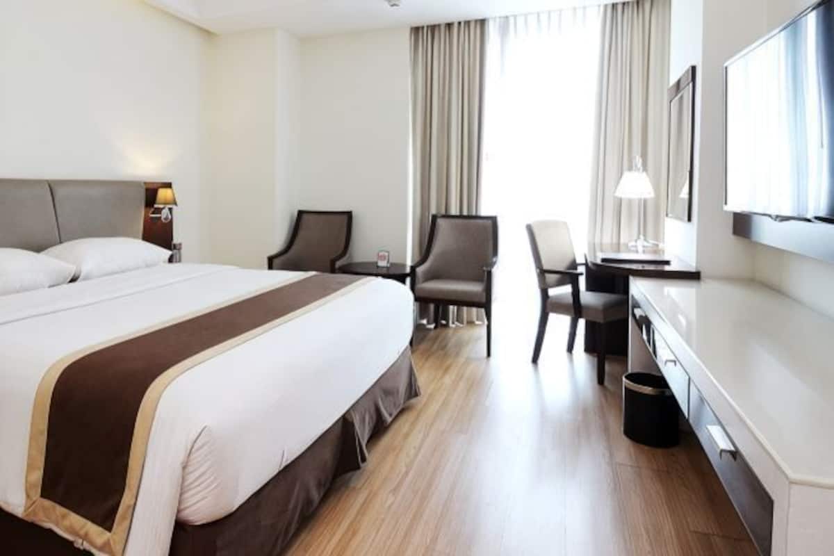 A hotel room with a large bed, a desk, two chairs, a TV, and a window with curtains.