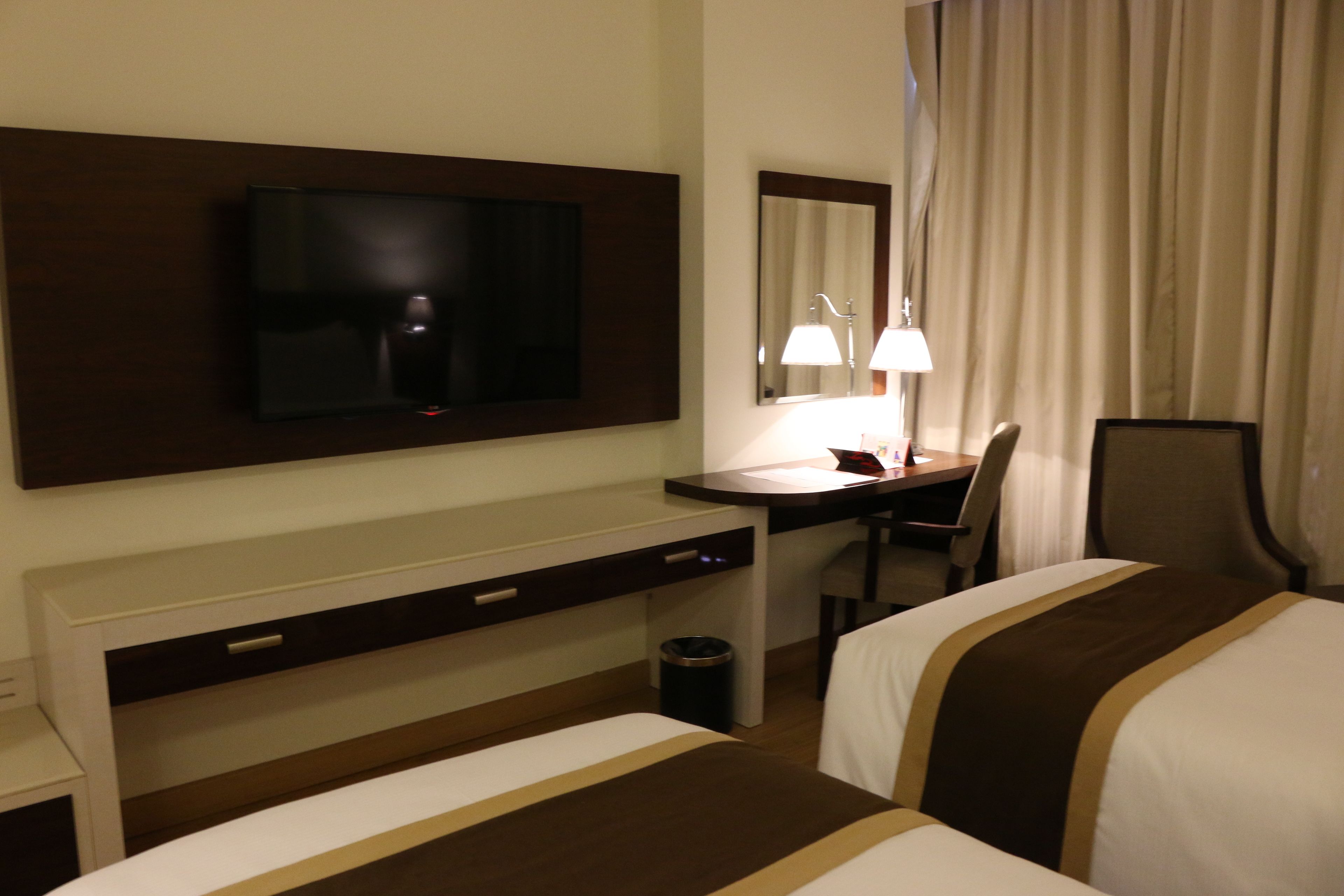 A hotel room with a TV mounted on the wall, a desk with a chair, a bedside table, and a bed with a striped bedspread.