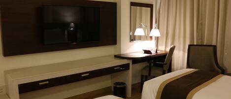 Habitación de hotel con televisión montada en la pared, escritorio con silla, mesita de noche y cama con cubrecama a rayas.
