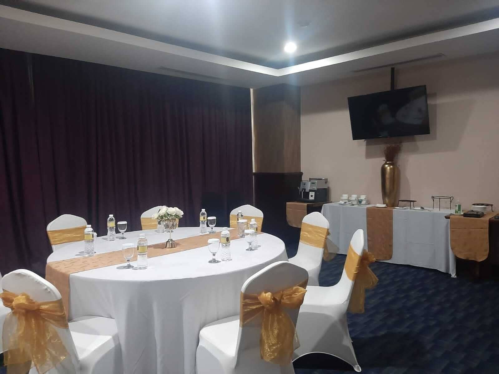 A conference room with round tables, white chairs with gold sashes, a large screen, and a stage with curtains.
