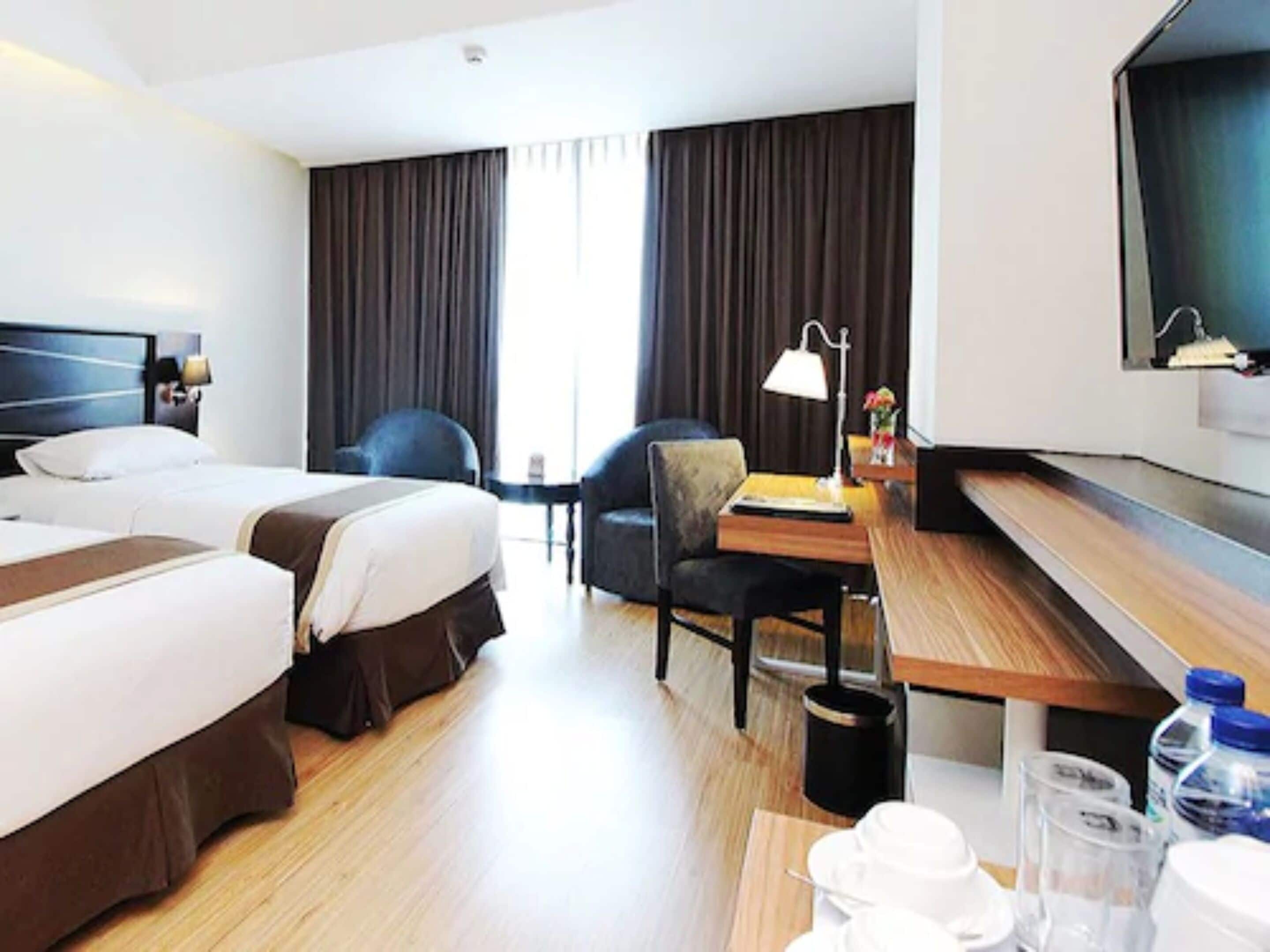 Ein Hotelzimmer mit zwei Betten, einem Schreibtisch mit Lampe, einem Sessel, einem an der Wand befestigten Fernseher und einem Fenster mit Vorhängen.