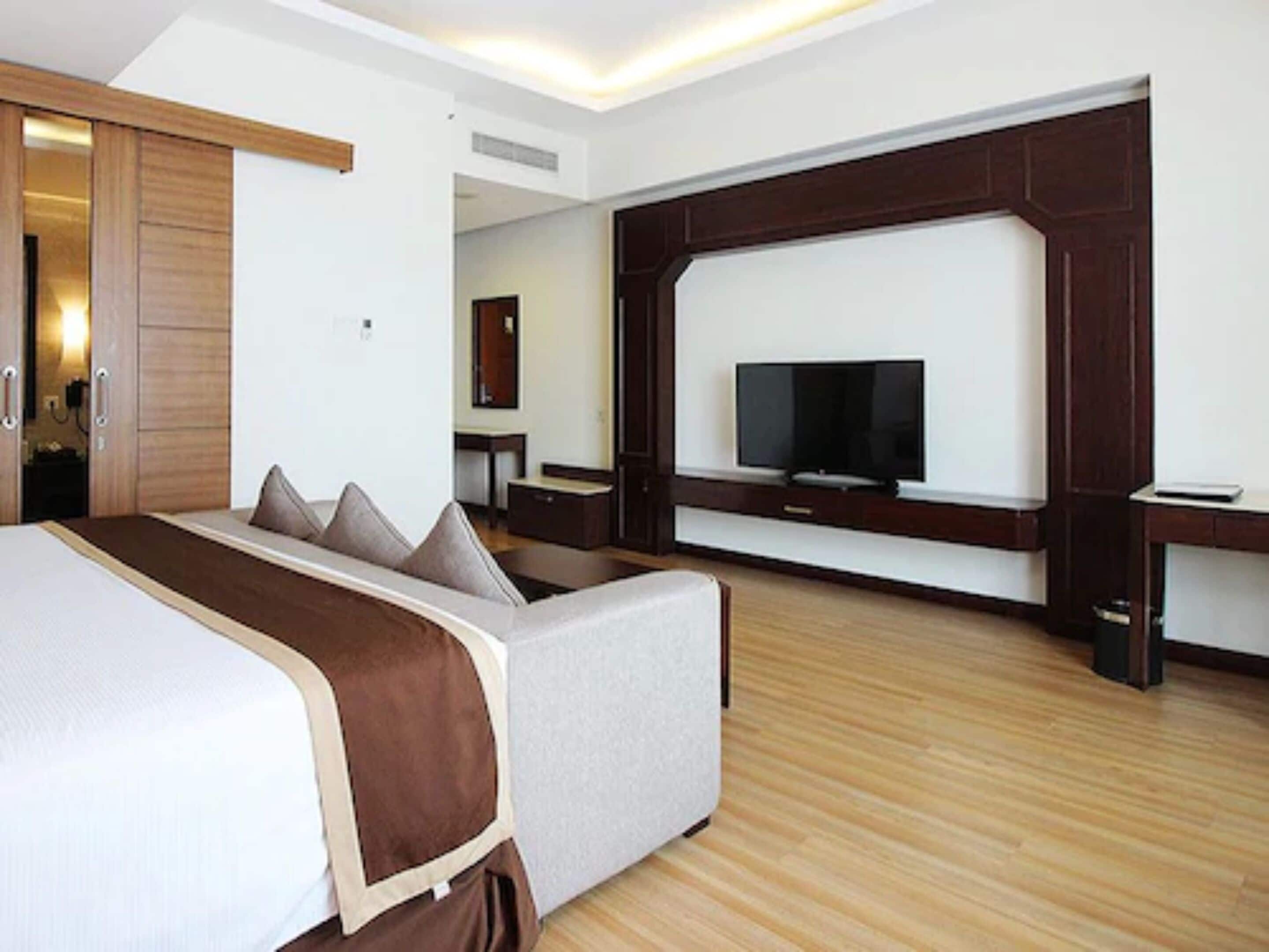 A modern hotel room with a large bed, a seating area, a TV mounted on the wall, and a wooden floor.