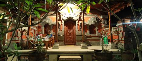 Un bâtiment traditionnel avec une porte en bois au centre, entouré de plantes en pot et de marches menant à l’entrée.