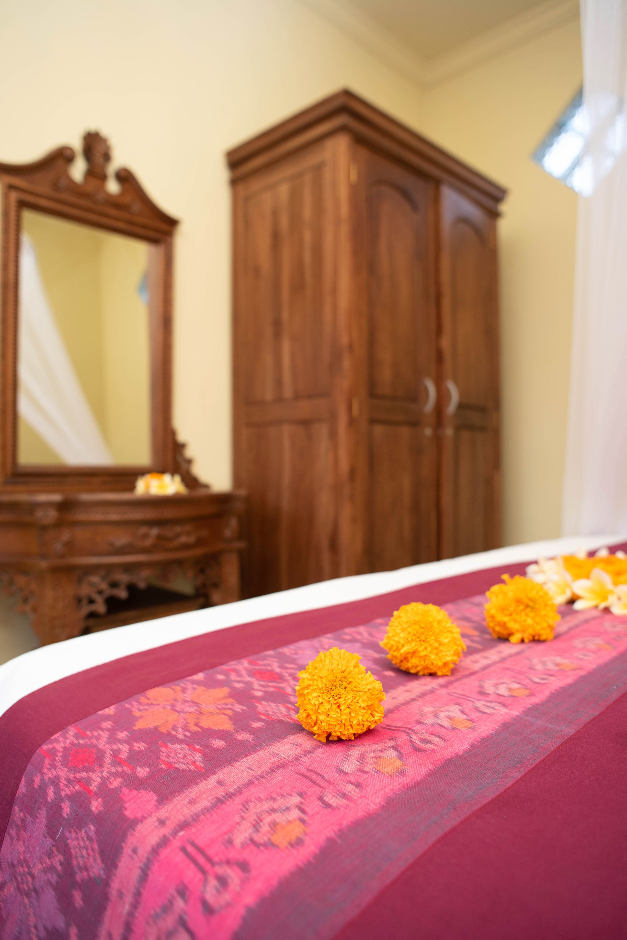 A bed with a pink and orange patterned bedspread and three yellow flowers. There is a wooden wardrobe and a mirror in the background.