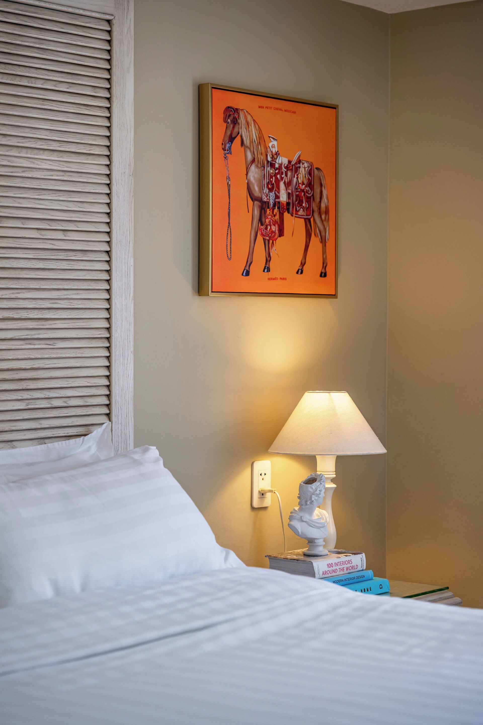 A hotel room with a bed, a nightstand with a lamp, a book, and a framed artwork on the wall.