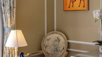 A corner room with a chair, a table with a lamp, a framed artwork, and a vase with flowers.