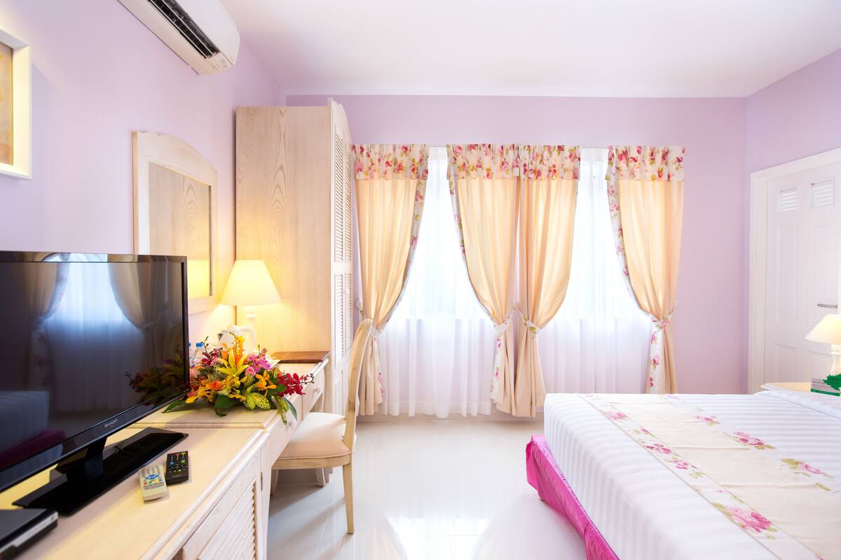 A hotel room with a flat-screen TV, a desk with a lamp, a chair, a bed with floral bedding, and a window with curtains.