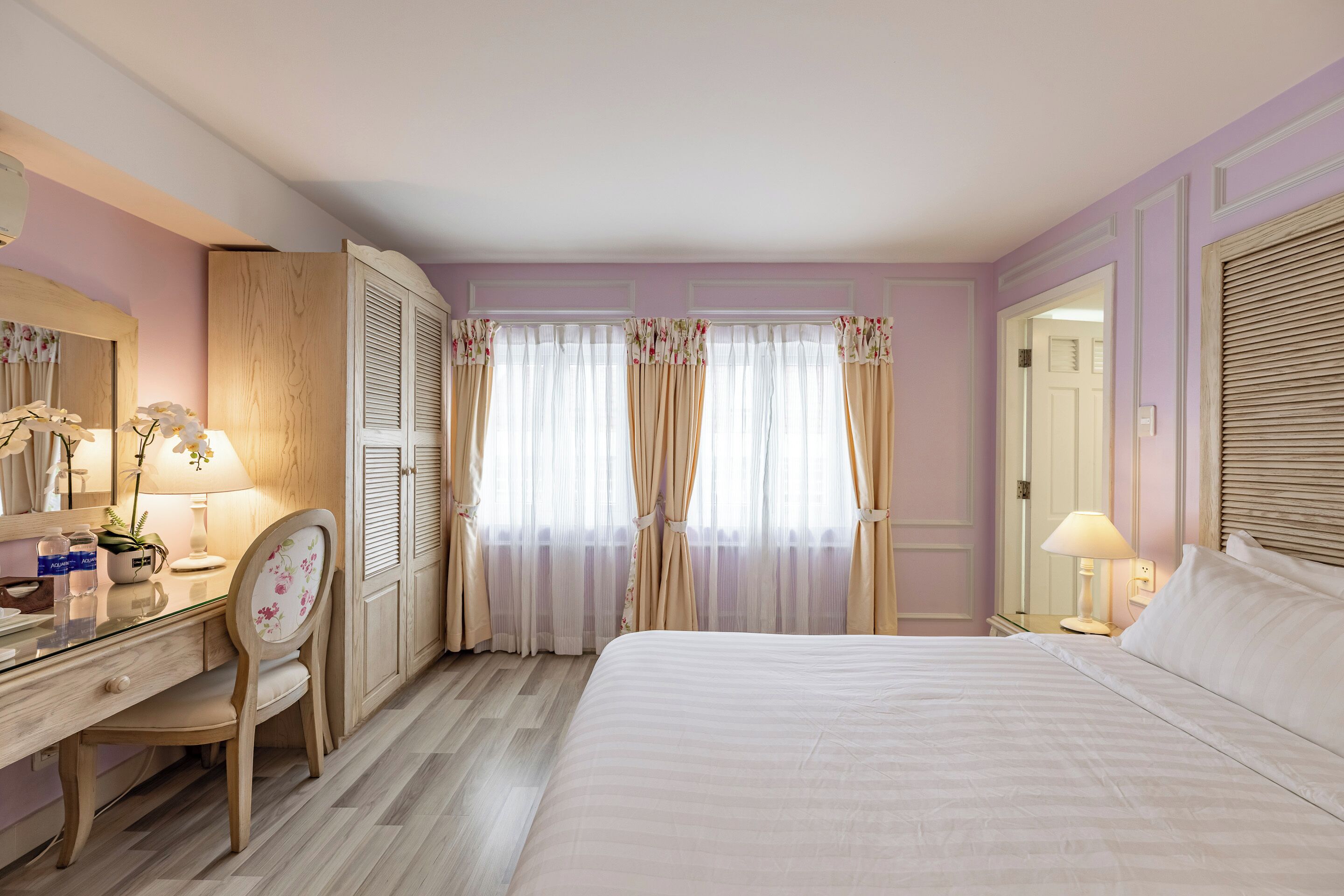 A hotel room with a bed, a desk with a chair, a mirror, and a window with curtains.